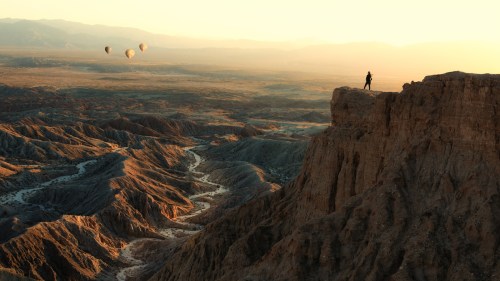 A lone figure stands on a cliff overlooking a vast desert landscape at sunset, with hot air balloons in the distance.