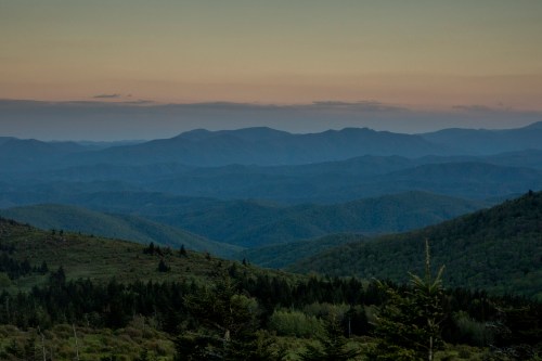 A serene mountain landscape at dusk, featuring rolling hills and a gradient sky transitioning from blue to soft orange.
