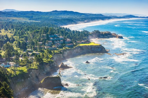 A scenic coastal view featuring cliffs, lush greenery, and waves crashing against the shore under a clear blue sky.