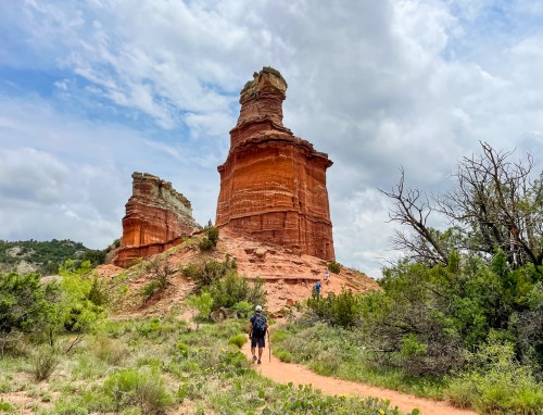 A hiker walks along a trail near towering red rock formations under a cloudy sky. Lush greenery surrounds the path.