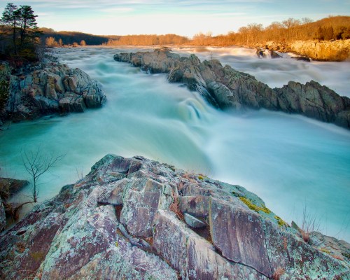 A serene river flows over rocky terrain, surrounded by trees and a tranquil landscape under a clear sky.