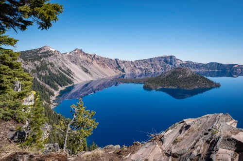 A serene view of Crater Lake, featuring clear blue water, a small island, and surrounding mountains under a clear sky.