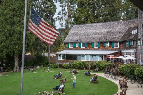A scenic view of a lodge with an American flag, surrounded by green grass and people relaxing on benches.