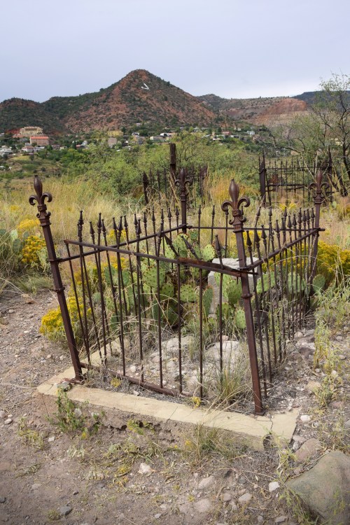 An old, ornate iron fence surrounds a grave in a desert landscape with mountains in the background.