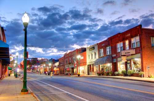 A quiet street lined with brick buildings and shops, illuminated by streetlights under a cloudy evening sky.