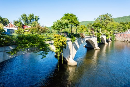 A stone bridge arches over a calm river, surrounded by lush greenery and blue skies.