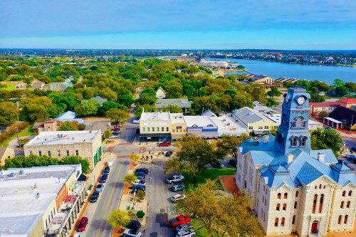 Aerial view of a town with historic buildings, green trees, and a river in the background under a clear blue sky.