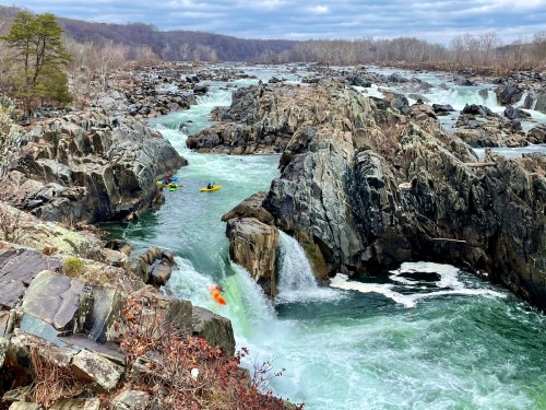 A scenic view of rocky rapids with kayakers navigating the turbulent waters under a cloudy sky.