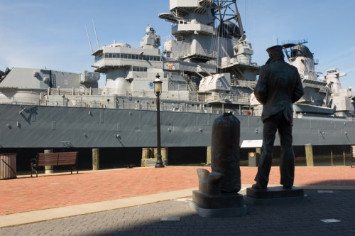 A statue of a sailor stands in front of a large battleship, with benches and a lamppost nearby.