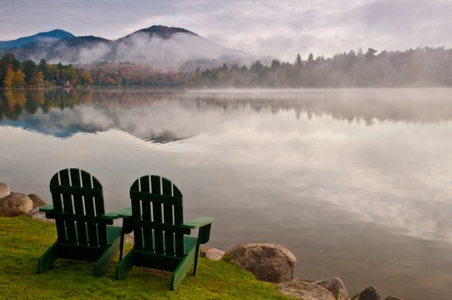Two green Adirondack chairs sit by a calm lake, surrounded by misty mountains and autumn foliage.
