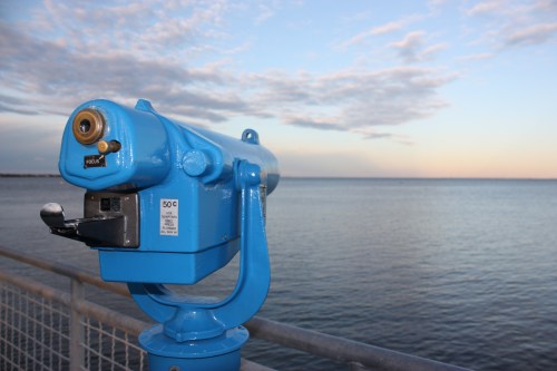 A blue telescope mounted on a railing, overlooking a calm body of water under a cloudy sky at sunset.