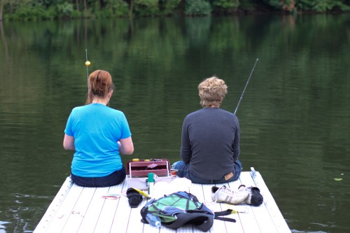 Two people sit on a dock, fishing by a calm lake surrounded by trees, with fishing gear and a tackle box nearby.