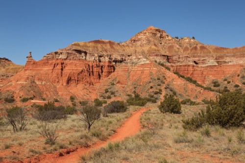 A winding dirt path leads through red rock formations and sparse vegetation under a clear blue sky.