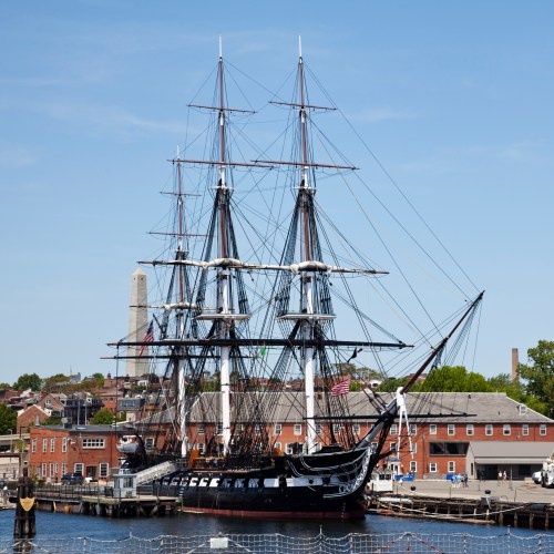 A historic tall ship docked at a harbor, with buildings and a monument visible in the background under a clear blue sky.