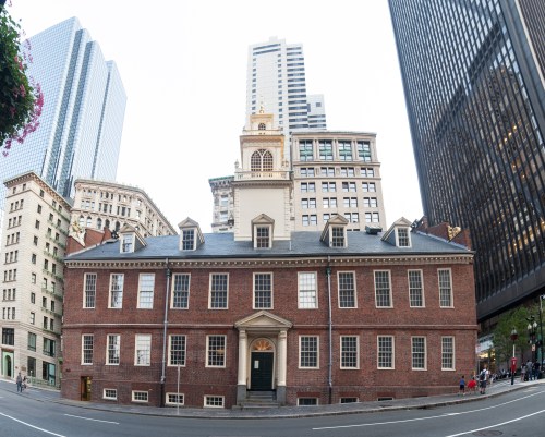 Historic brick building with a clock tower, surrounded by modern skyscrapers in an urban setting.