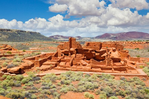 Ancient red rock ruins surrounded by green vegetation and mountains under a blue sky with fluffy clouds.