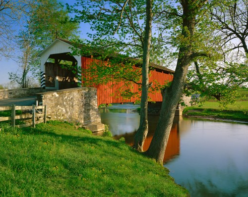 A scenic view of a covered bridge over a calm river, surrounded by lush greenery and trees.