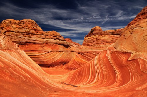 Vibrant, swirling sandstone formations in a desert landscape under a blue sky with wispy clouds.