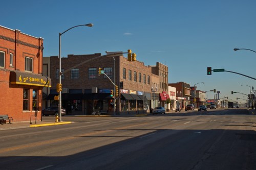 A quiet street scene featuring brick buildings, shops, and traffic lights under a clear blue sky.