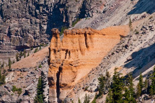 A rocky cliff formation with orange and gray layers, surrounded by green trees and rugged terrain.