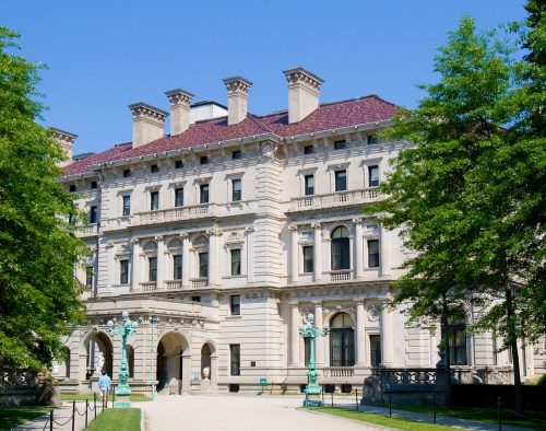 A grand historic mansion with ornate architecture, surrounded by green trees and a clear blue sky.