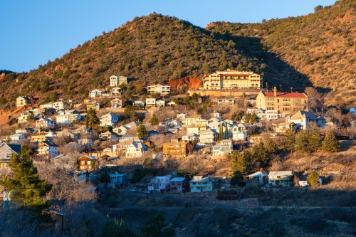 A hillside community with various houses and buildings, surrounded by mountains and trees, bathed in warm sunlight.