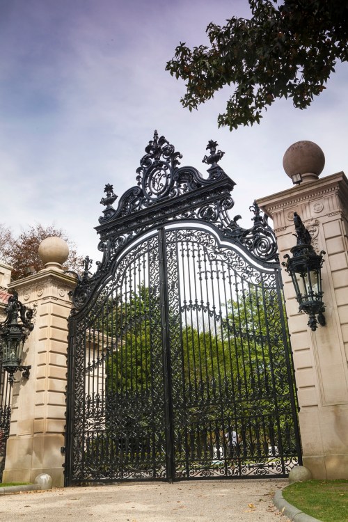 Ornate black wrought iron gates with decorative details, flanked by stone pillars and lanterns, set against a cloudy sky.