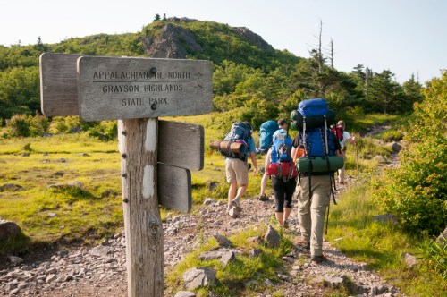 Hikers with backpacks walking on a trail near a wooden sign for Grayson Highlands State Park.
