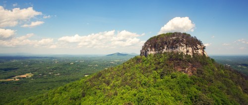 A panoramic view of a lush green mountain with a rocky summit under a blue sky with scattered clouds.