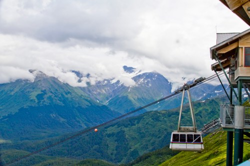 A gondola lifts from a mountain station, surrounded by lush green hills and cloudy peaks in the background.