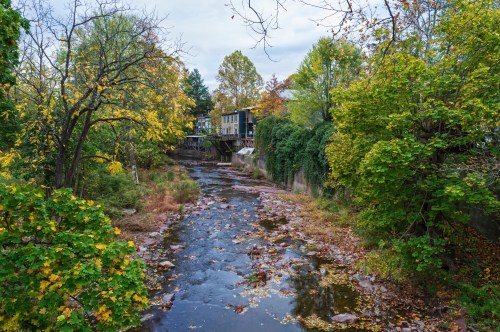 A serene creek surrounded by colorful autumn foliage and a house in the background under a cloudy sky.