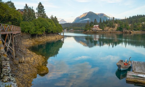 Serene waterfront scene with a calm bay, lush trees, mountains in the background, and a small boat docked nearby.