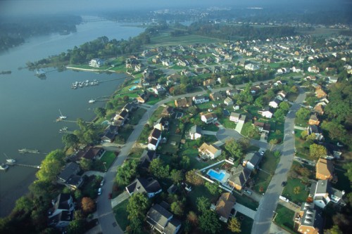 Aerial view of a suburban neighborhood by a river, featuring houses, greenery, and a few boats on the water.