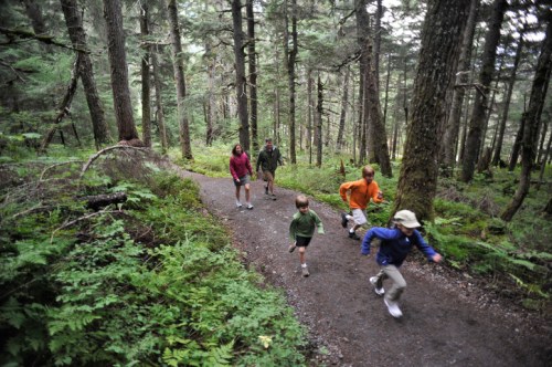 A group of five people, including children, walking and running on a forest trail surrounded by tall trees and greenery.