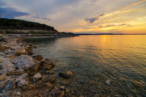 A serene lakeside view at sunset, with calm waters reflecting the colorful sky and rocky shoreline.