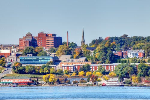 A scenic view of a riverside city with colorful autumn trees, buildings, and a clear blue sky.