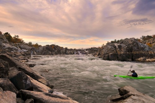 A kayaker navigates a rocky river with swirling waters under a colorful sky at sunset.