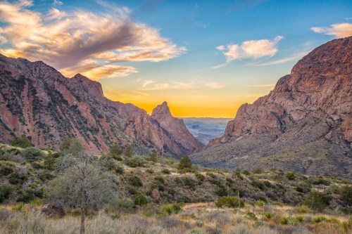 A scenic view of mountains at sunset, with vibrant clouds and a valley filled with greenery in the foreground.