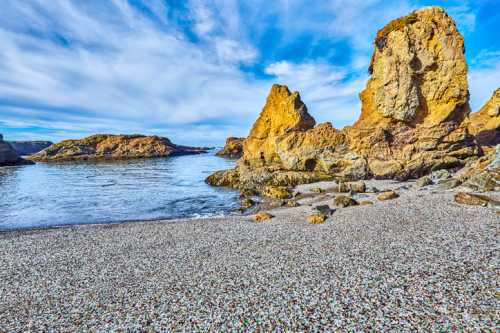 Rocky coastline with golden cliffs and calm blue water under a partly cloudy sky. Pebble beach in the foreground.