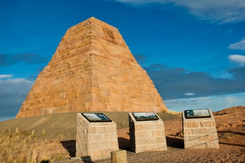 A stone pyramid structure with informational plaques in the foreground against a blue sky.