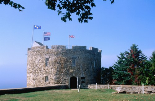 A stone tower with flags flying, surrounded by green grass and trees under a clear blue sky.