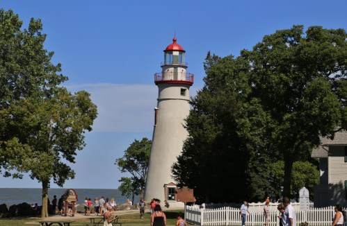 A tall lighthouse surrounded by trees, with people walking nearby and a clear blue sky above.