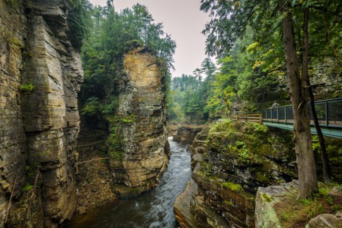 A serene river flows through a rocky gorge, surrounded by lush greenery and towering cliffs. A pathway runs alongside.