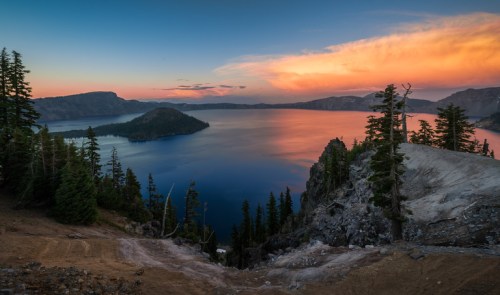 A serene view of Crater Lake at sunset, with vibrant colors reflecting on the water and trees framing the scene.