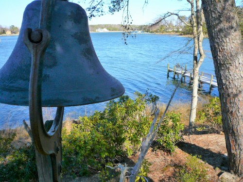 A bell hangs near a tranquil waterfront scene with a dock and trees by the water's edge.