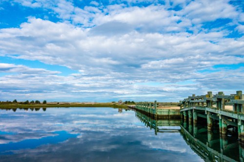 A serene waterfront scene with a wooden dock reflecting in calm water under a blue sky with fluffy clouds.
