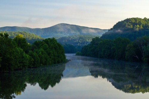 A serene river reflects lush green trees and misty mountains under a soft morning sky.