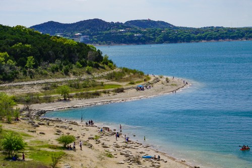 A scenic view of a sandy shoreline with people enjoying the beach, surrounded by green hills and a calm blue lake.