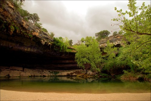 A serene natural scene featuring a calm pool surrounded by lush greenery and rocky cliffs under a cloudy sky.