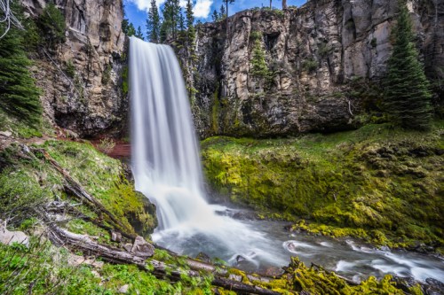 A serene waterfall cascades down rocky cliffs, surrounded by lush greenery and tall trees under a blue sky.